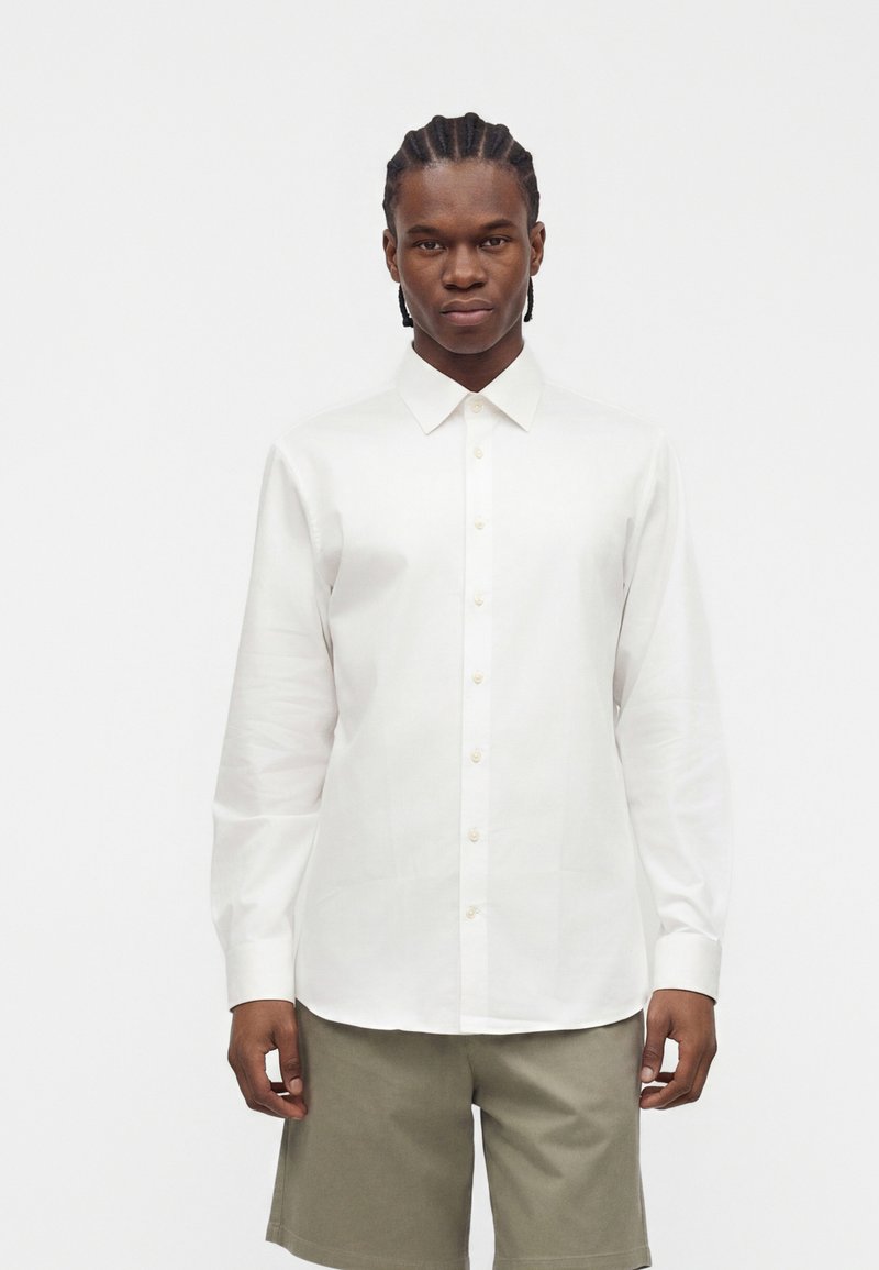 Hombre con el cabello trenzado que lleva una camisa blanca de manga larga con botones y pantalones cortos beige, de pie frente a un fondo blanco liso.