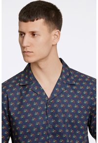 Korte mouwen shirt van marineblauwe stof met een herhalend patroon van kleine fietsen in groen, rood en geel. Met een ingesneden kraag en knoopsluiting.