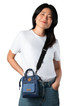 Junge Frau mit langen dunklen Haaren trägt ein weißes T-Shirt und Jeans und hat eine kleine blaue Umhängetasche mit einer vorderen Reißverschlusstasche dabei.