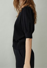 Žena s dlouhými hnědými vlasy, která má na sobě černý svetr s krátkým rukávem a černé kalhoty, před jednobarevným světle šedým pozadím.