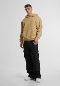 Felpa con cappuccio beige con tasca frontale, abbinata a pantaloni cargo neri con più tasche e sneakers bianche.