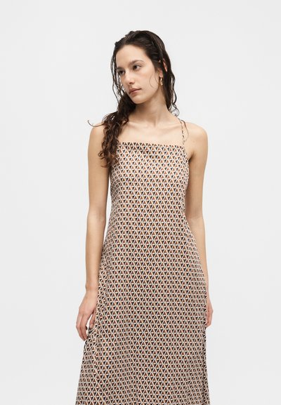 Νέα γυναίκα με βρεγμένα, σγουρά μαλλιά φοράει αμάνικο, μάξι φόρεμα καφέ και λευκού μοτίβου, στέκεται μπροστά σε απλό φωτεινό φόντο.