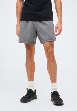 Mand iført grå Nike-shorts, sorte sneakers, hvide crew-sokker og en sort T-shirt, stående på en ensfarvet lys baggrund.