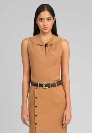 Femme portant une robe camel sans manches avec une ceinture marron foncé et des détails de boutons décoratifs sur le côté gauche, debout devant un fond gris.