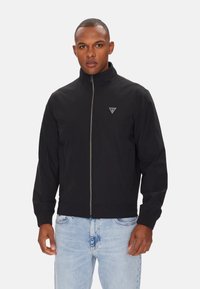 Giacca nera con cerniera e collo alto, polsini elasticizzati e un logo triangolare sul petto. Indossata sopra jeans azzurri.