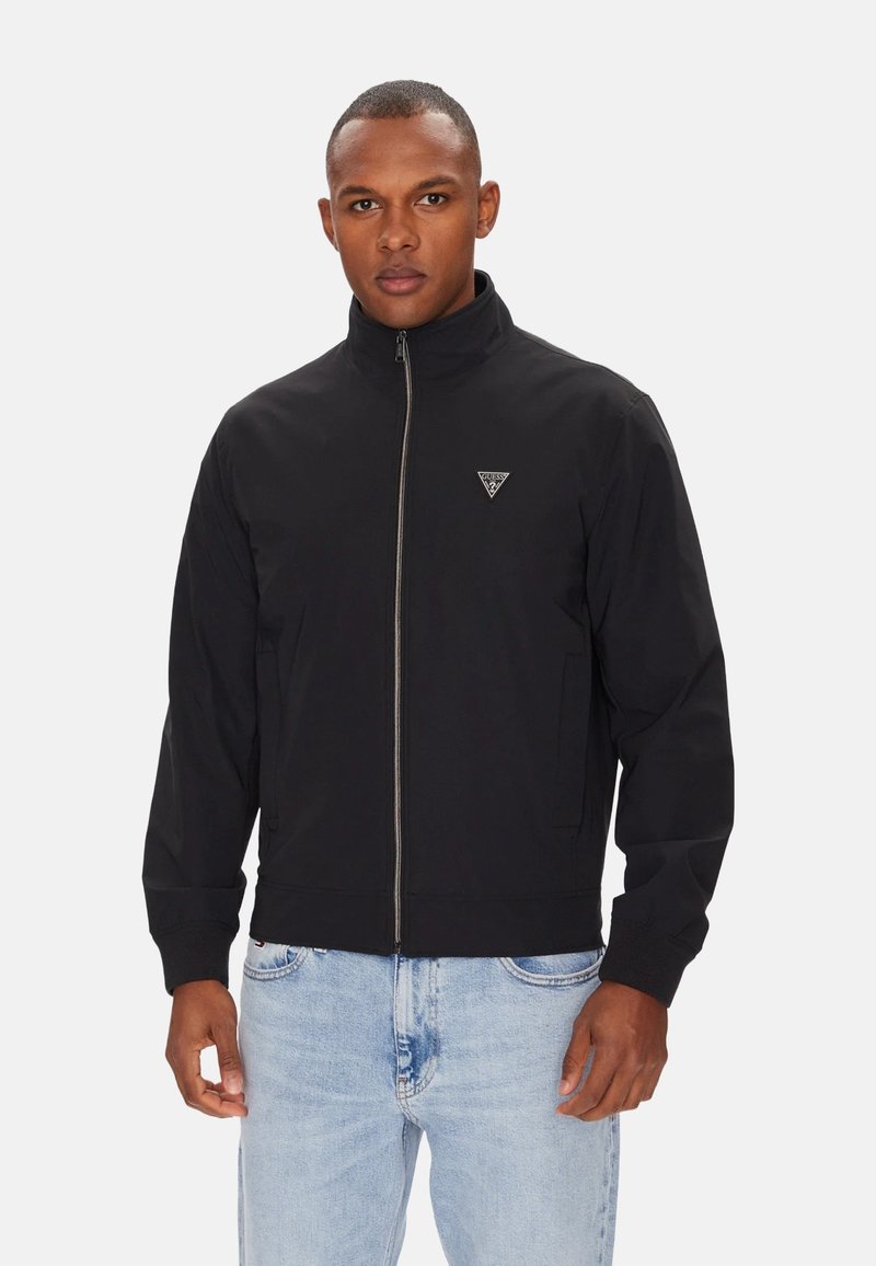 Giacca nera con cerniera e collo alto, polsini elasticizzati e un logo triangolare sul petto. Indossata sopra jeans azzurri.