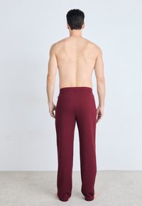 Bordeaux kleurige relaxte broek met een zachte textuur. Elastische tailleband, wijde pijpen en geen zichtbare patronen. Draagt door een persoon van achteren.