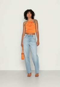 Mulher com cabelo cacheado, vestindo um top halter laranja, jeans de cintura alta azul clara, saltos laranjas, segurando uma bolsa laranja, de pé contra uma parede simples.