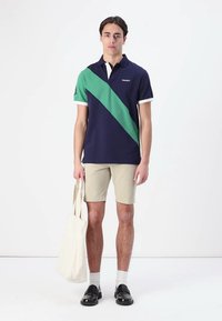 Polo skjorte i marineblå med en grønn diagonallinje, hvit krage og korte ermer. Kombinert med beige shorts og svarte slip-on sko.