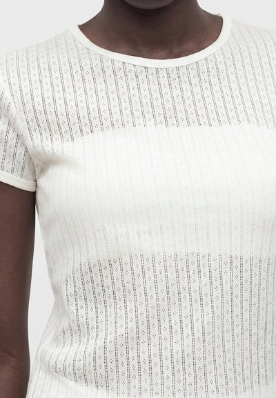 Άτομο με σκουρόχρωμο δέρμα που φοράει λευκό, κοντομάνικο, ημιδιαφανές πλεκτό τοπ με κάθετα σχέδια από μικρές οπές και στρογγυλή λαιμόκοψη.