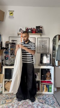 Witte kleding vastgehouden door een persoon, met een zwart-wit gestreept polo, losse zwarte broek en een patroonkleed eronder. Verschillende items op de achtergrond.