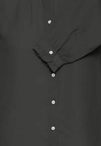 Blouse noire à manches longues avec des boutons blancs sur le devant et sur le poignet froncé de la manche.