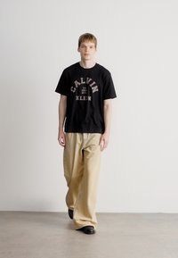 Črn majica s potiskom "CALVIN KLEIN" v vijoličasti in beli barvi, v kombinaciji z ohlapnimi bež hlačami in črnimi čevlji. Gladek material, vsakdanji kroj.