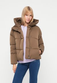 Casaco puffer castanho com capuz, apresentando um design matelassado e fecho na frente. Usado sobre um suéter de cor roxa clara e jeans azuis.