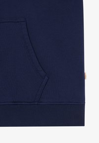Gros plan sur la poche d'un sweat-shirt bleu marine avec des coutures visibles et une petite étiquette en tissu sur le côté droit.