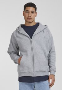 Gray zip-up hoodie with a soft texture, adjustable drawstring hood, and kangaroo pockets, layered over a navy shirt. Casual fit.