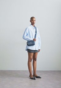 Cămașă în dungi albastre, fustă din denim și geantă crossbody din material închis. Modelul poartă pantofi negri cu accente decorative. Fundal minimalist.