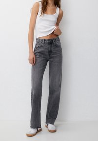 Mujer con jeans rectos de talle alto grises, camiseta blanca levantada para revelar el abdomen, y zapatillas blancas con suelas de goma.