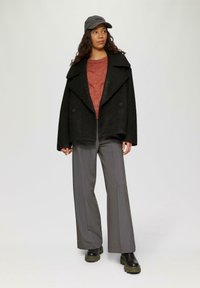 Zwarte textuur coat met brede schouders, rode gebreide trui, grijze wijde broek, zwarte schoenen met dikke zool en een grijze pet.
