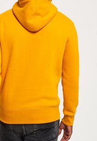 Sudadera con capucha amarilla de acabado texturizado. Presenta un dobladillo y puños ajustados. La vista trasera muestra el diseño de la capucha y sin logotipos visibles.