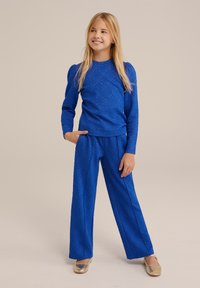 Leende tjej med långt blont hår iklädd en strukturerad blå långärmad topp och matchande vida byxor, guldskor, poserande mot en neutral bakgrund.