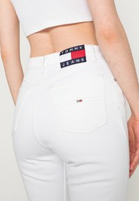 Jeans de mezclilla blanca con bolsillos traseros, con una etiqueta de marca en azul marino con acentos en rojo y blanco. Textura suave, diseño de pierna recta.