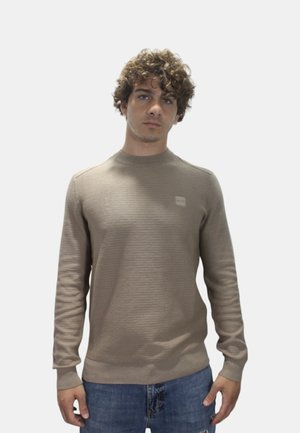 Felpa a maglia marrone chiaro con scollo rotondo, polsini a coste e un piccolo patch con logo sul petto, abbinata a jeans di denim blu.