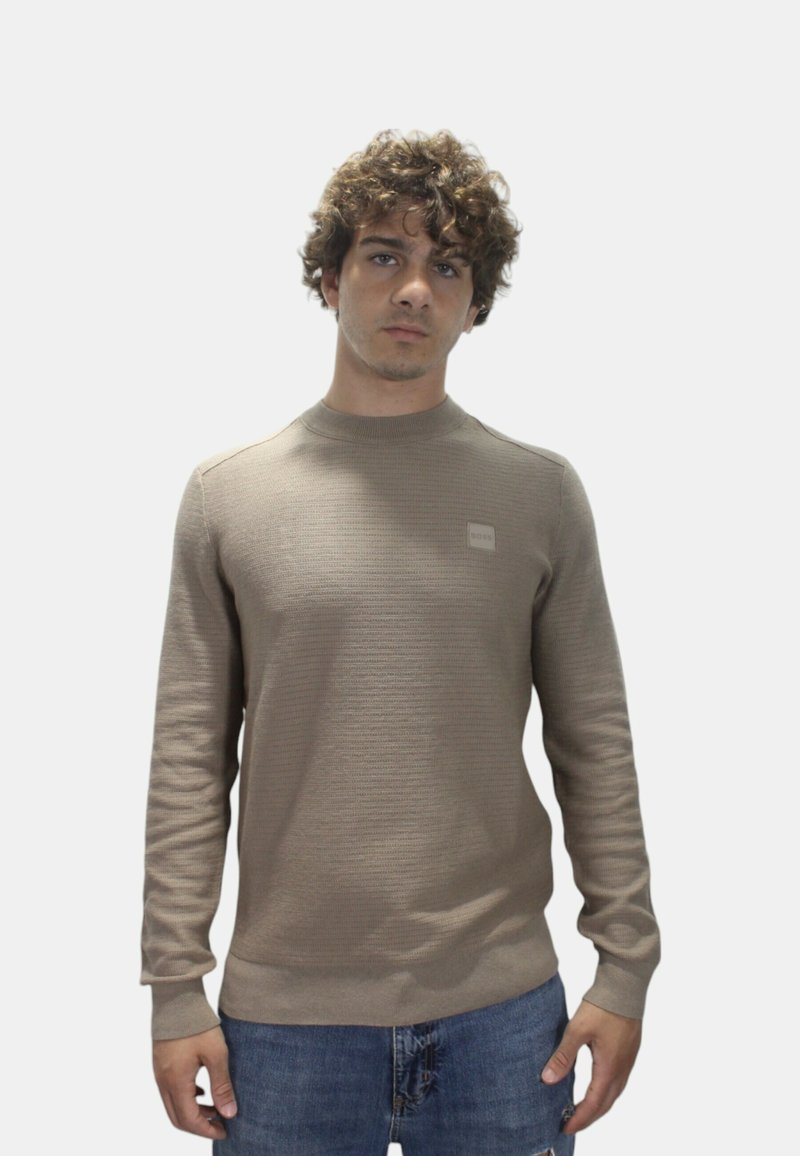 Felpa a maglia marrone chiaro con scollo rotondo, polsini a coste e un piccolo patch con logo sul petto, abbinata a jeans di denim blu.