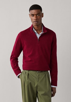 Homme portant un pull bordeaux à zip quart sur une chemise rayée, associé à un pantalon plissé vert olive et une ceinture marron, debout contre un mur uni.