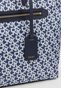 Námořnicky modrá a bílá taška přes rameno s opakujícím se vzorem loga DKNY. Hladký texturovaný povrch se zlatým zipem a zavazadlovou visačkou s logem značky.