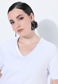 T-shirt bianca a V in tessuto a costine. Orecchini neri rettangolari grandi con accenti argentati. Capelli neri corti e lisci.