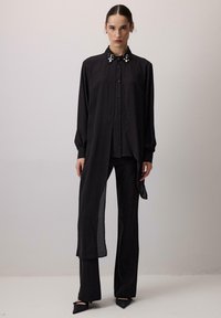 Czarna bluzka z długim rękawem z ozdobnym kołnierzykiem i zdobieniami, w połączeniu z dopasowanymi czarnymi spodniami i czółenkami z wystającym noskiem.