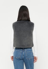 Femme avec des cheveux sombres et raides porte un gilet en fourrure gris sur un haut à manches longues blanc et un jean en denim bleu, photographiée de dos.