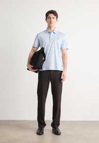 Polo de cor azul clara com mangas curtas, calças pretas e uma mala preta. Tecido liso com um pequeno logótipo na camisa. Sapatos pretos formais.