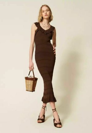 Donna che indossa un abito midi marrone senza maniche con bordi arricciati, tiene una borsa di vimini e indossa tacchi marroni stringati.