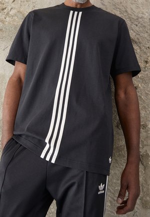 Mann trägt ein schwarzes Adidas-Outfit mit drei vertikalen weißen Streifen auf dem T-Shirt und Adidas-Logo auf der Hose vor einer strukturierten Wand.