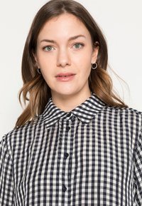 Camisa xadrez em preto e branco, feita de um tecido leve, com gola, fecho de botões à frente e mangas curtas.