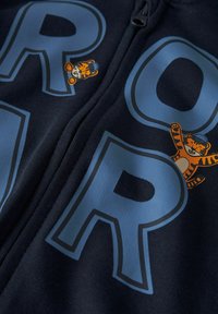Sudadera con cremallera azul navy, con las letras grandes en azul claro "R" y "O", y un pequeño gráfico de un tigre naranja en el lado izquierdo cerca de la cremallera.