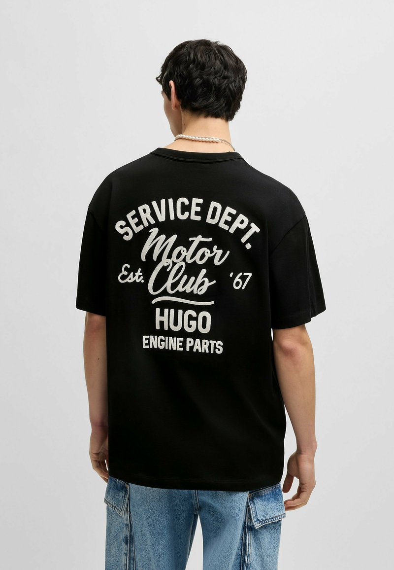 Zwarte katoenen t-shirt met een oversized pasvorm. Kenmerkt witte grafische tekst op de achterkant: "SERVICE DEPT. Motor Club '67 HUGO ENGINE PARTS."