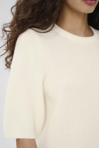 Maglione di colore crema a maglia con maniche corte e leggermente a sbuffo, e scollatura rotonda. Texture morbida con una sensazione leggera e ariosa.