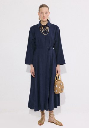 Žena v tmavomodrom dlhom rukávovom šatách s opaskom a vrstvenými zlatými náhrdelníkmi, držiaca malú béžovú kabelku s korálkami, nosiaca béžové šnurovacie sandále.