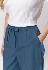 Plave lagane hlače s elastičnim pojasom, podesivim vezicama, bočnim džepovima i malim izvezenim detaljem na kuku.