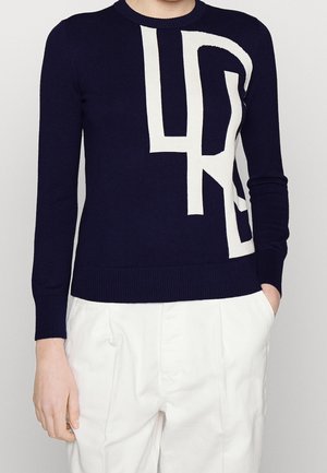 Personne portant un pull bleu marine avec un grand motif géométrique abstrait blanc et un pantalon blanc, les mains partiellement dans les poches.