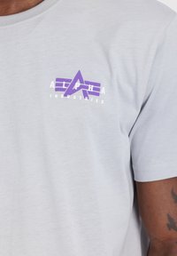 Hallgrå puuvillane särk, millel on rinnas lilla ja valge "Alpha Industries" logo. Lühikesed varrukad ja ümmargune kaelus.