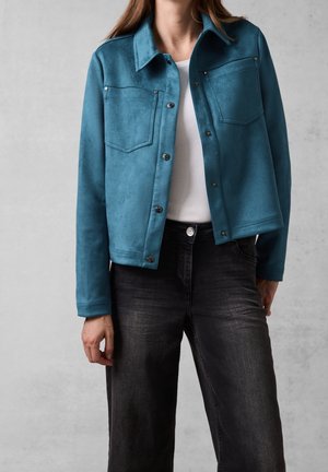 Kobieta w zamszowej kurtce na guziki w kolorze teal, na białym topie i czarnych dżinsach, stojąca na tle jednolitego jasnoszarego tła.