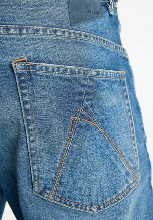 Kék denim farmernadrág hátsó zsebe narancssárga varrással és "CHASIN'" felirattal ellátott bőrcímkével.
