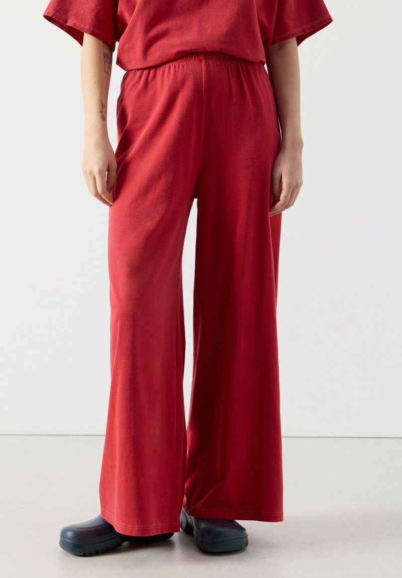 Osoba ubrana w luźne, wysokiego stanu, szerokie czerwone spodnie i czarne buty wsuwane, stojąca na tle jednolitego jasnego tła.