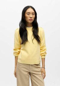 Žlutý svetr s žebrovaným výstřihem a nafouklými rukávy, v kombinaci s béžovými kalhotami, hladká textura, volnočasový design.
