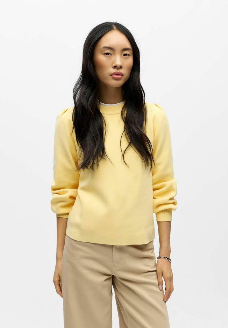 Žlutý svetr s žebrovaným výstřihem a nafouklými rukávy, v kombinaci s béžovými kalhotami, hladká textura, volnočasový design.