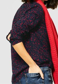 Mujer vistiendo una blusa azul marino con estampado floral rojo, una bufanda texturada roja y jeans azules con una mano en el bolsillo trasero.